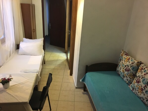 Una habitación pequeña y bien iluminada con una cama a la izquierda, una silla y un sofá a la derecha. Se ve una puerta que da a otra habitación y una pequeña mesa con un florero.