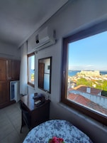 Un dormitorio con un ventanal grande, un escritorio, una silla, una cama con colcha estampada y vistas a un paisaje urbano costero.