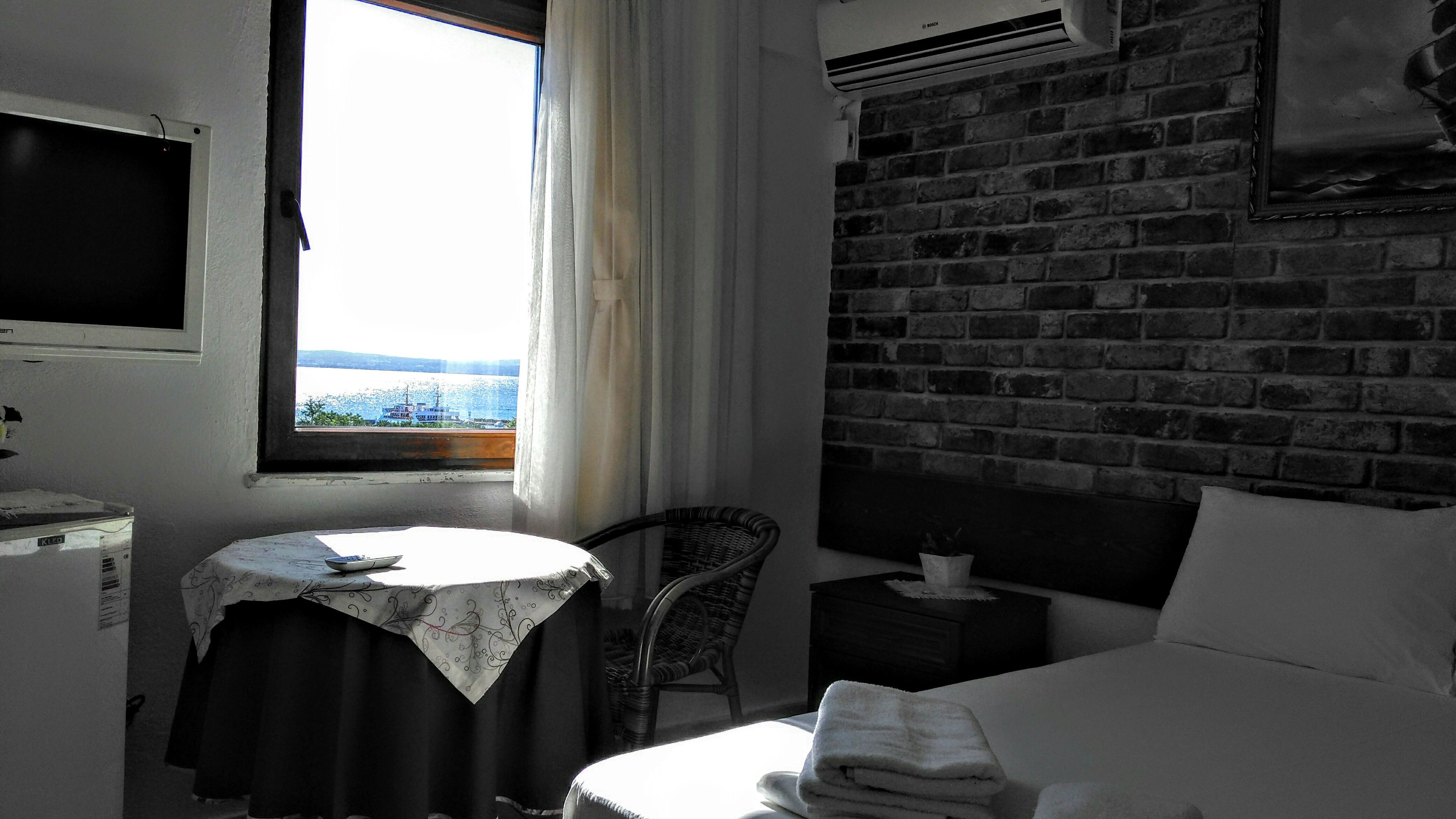 A hotel room with a bed, a small table with a remote control, a chair, a nightstand, and a view of the sea through a window.