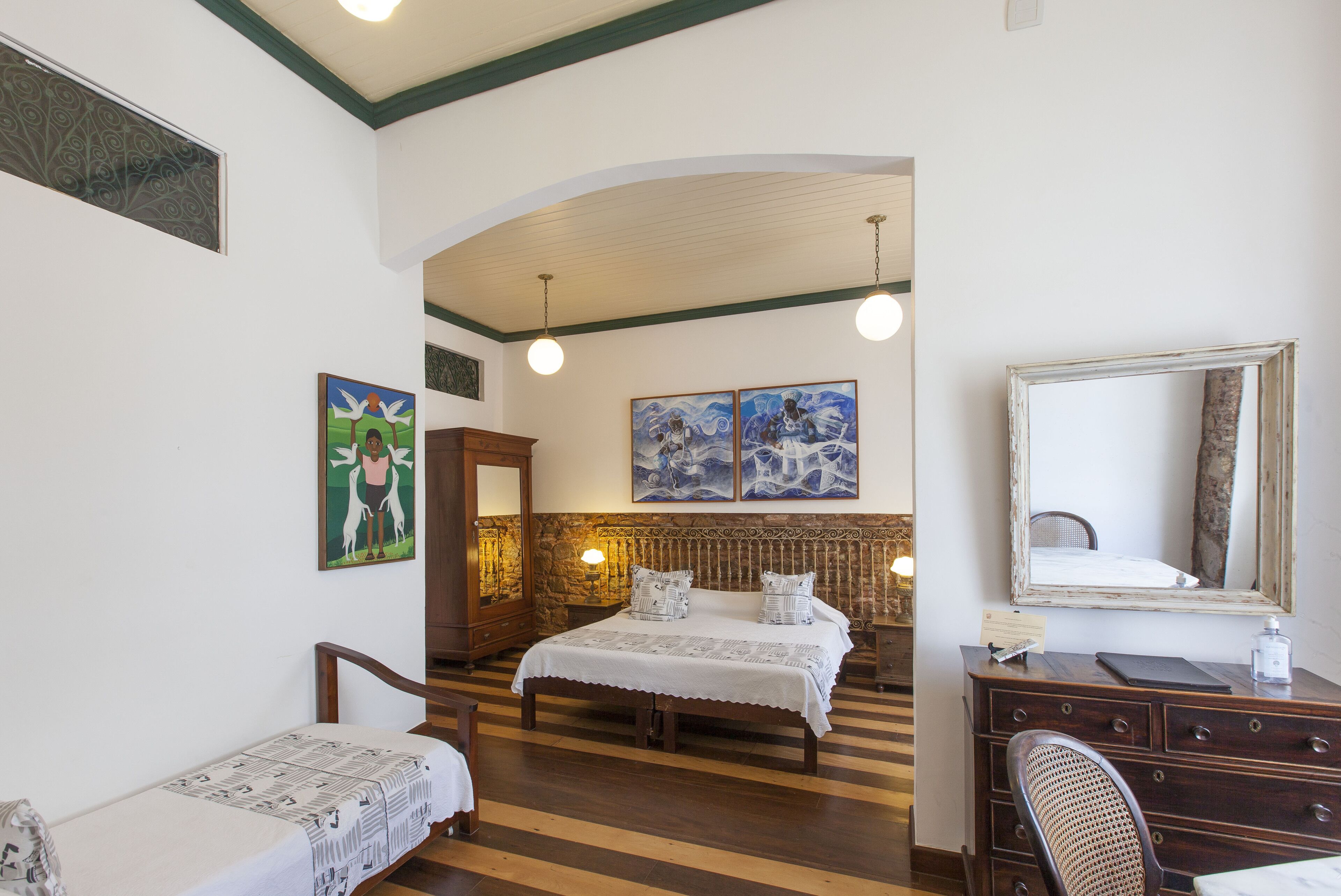 Un dormitorio con una cama de madera, un pequeño sofá, un aparador y un espejo. Hay cuadros en la pared y una ventana con un diseño decorativo.