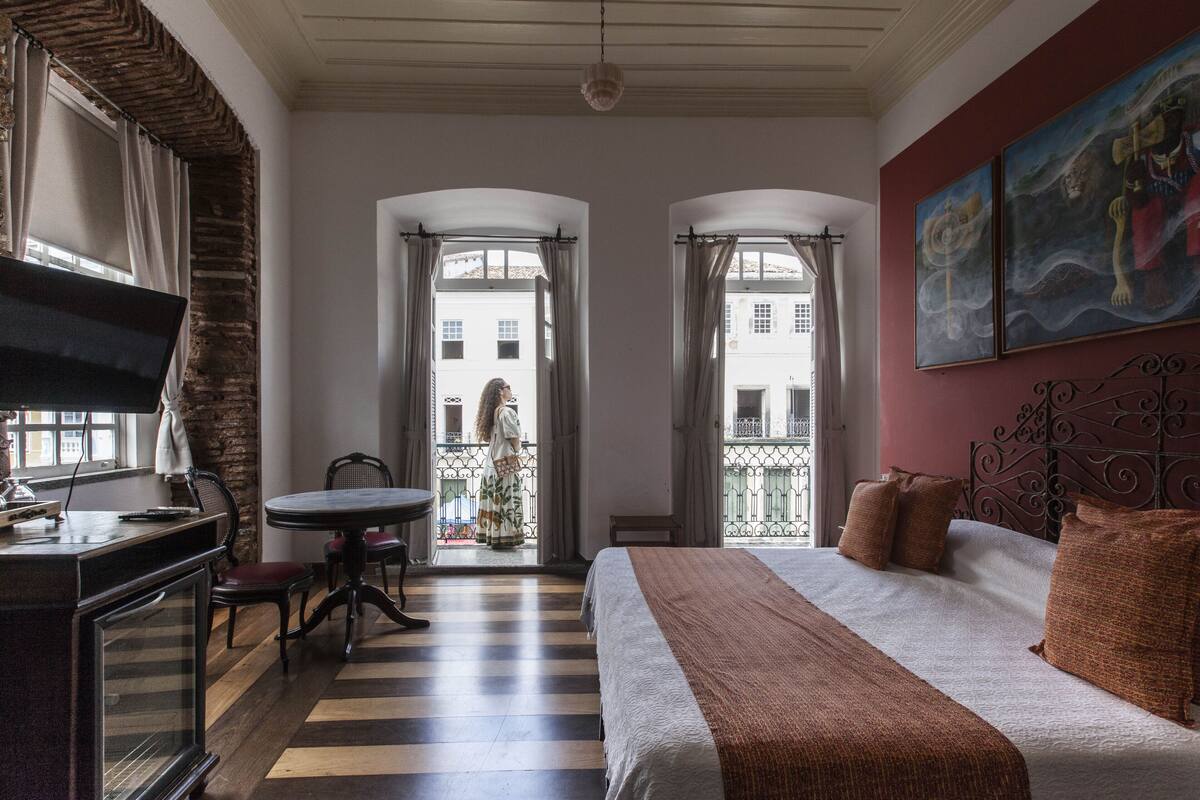 A hotel room with a bed, a desk, a chair, a table, and a view of the outside balcony.