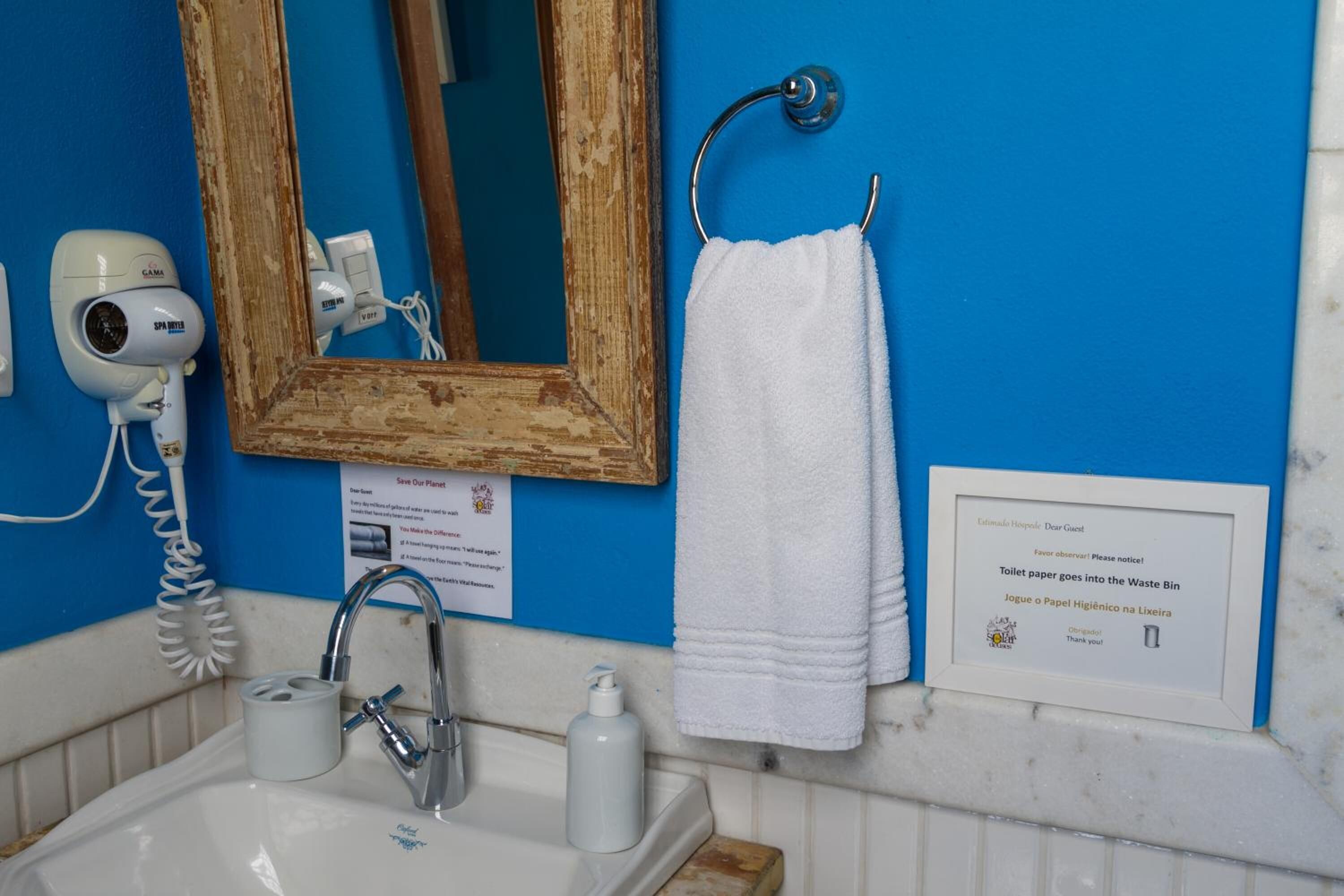 Un baño con lavamanos, espejo, toallero y un cartel que indica desechar el papel higiénico en el cubo de basura.