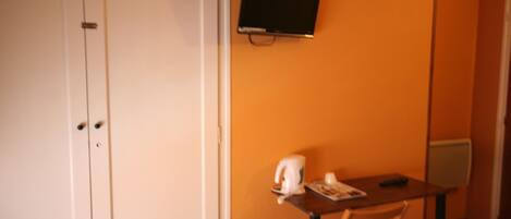 Een kleine kamer met een witte deur, een tafel met waterkoker en kopjes, een televisie aan de muur, en een radiator.