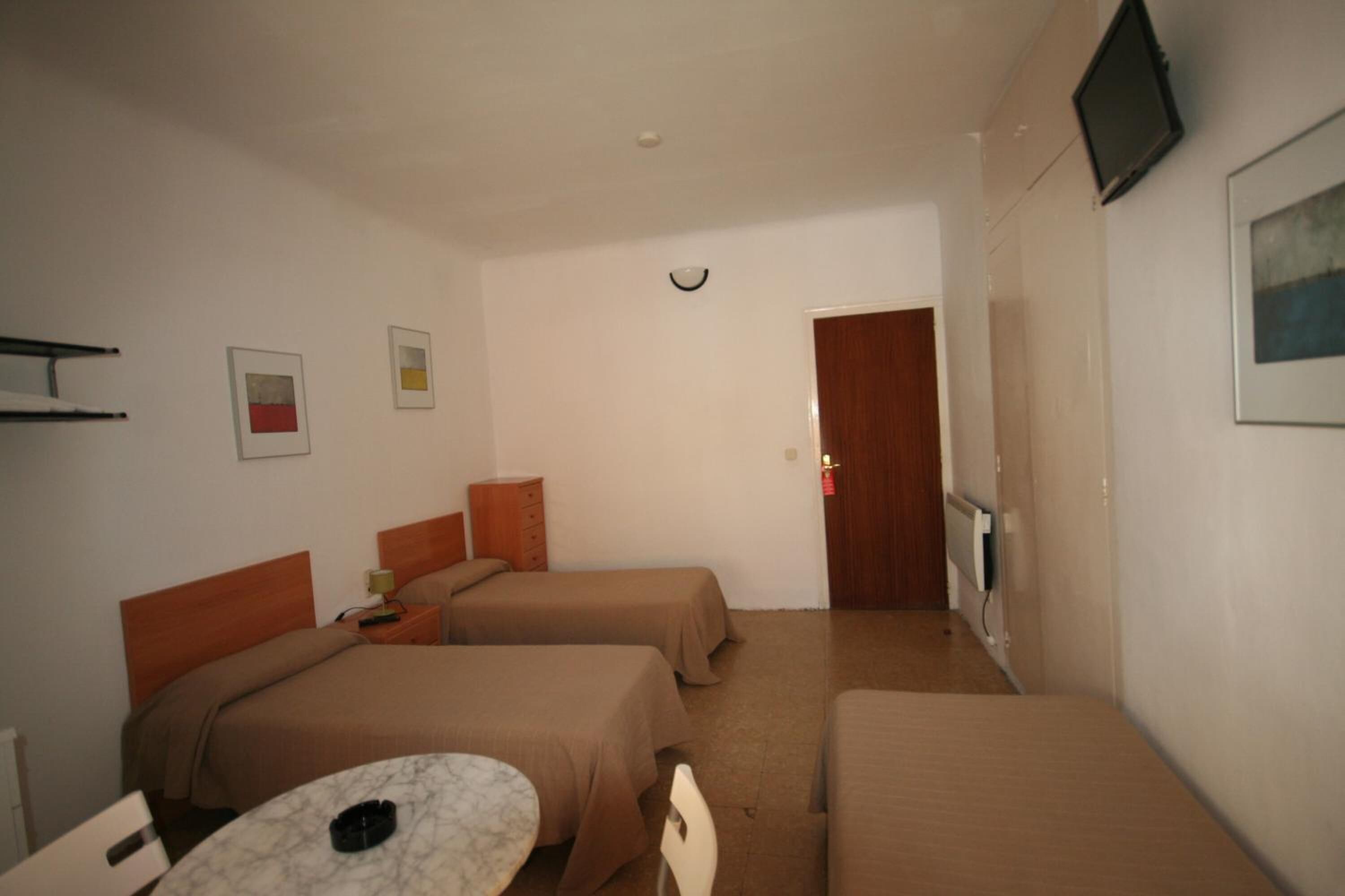 A hotel room with two single beds, a small table, a chair, a TV mounted on the wall, and a door leading to another room.