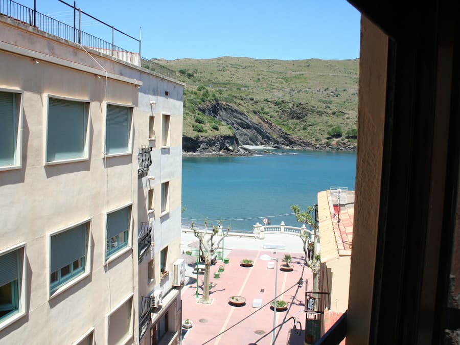 Vue d’un bâtiment situé sur la côte, doté de nombreuses fenêtres et de balcons, qui offre une vue sur une mer calme et un paysage vallonné.
