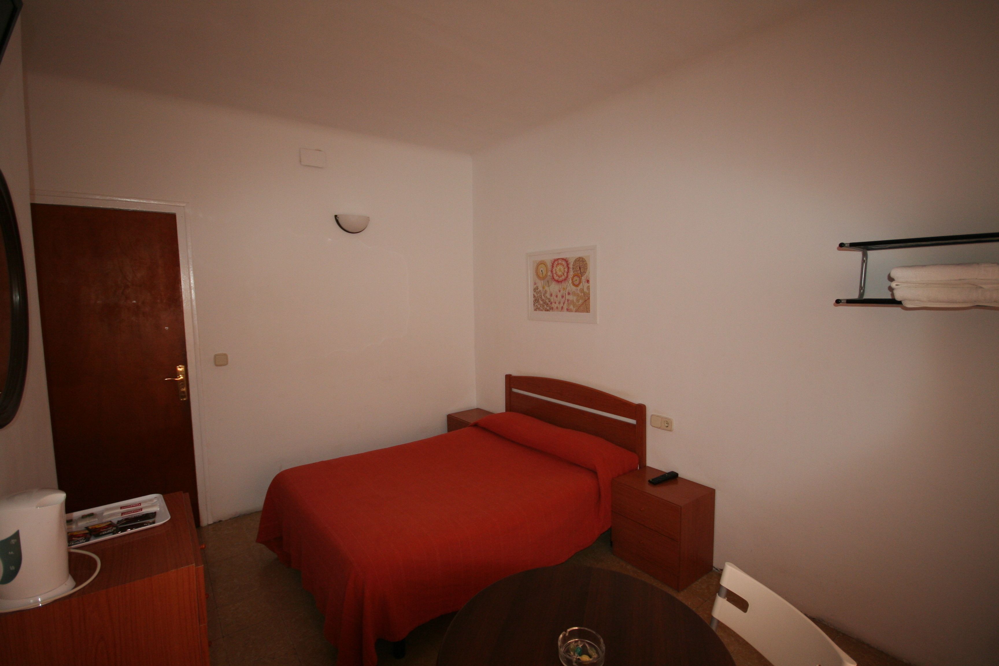 A bedroom with a single bed, a bedside table, a nightstand, a wooden dresser, a round table, and a wall-mounted shelf with towels.