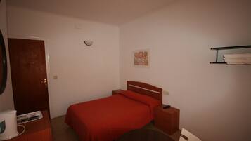 A bedroom with a single bed, a bedside table, a nightstand, a wooden dresser, a round table, and a wall-mounted shelf with towels.