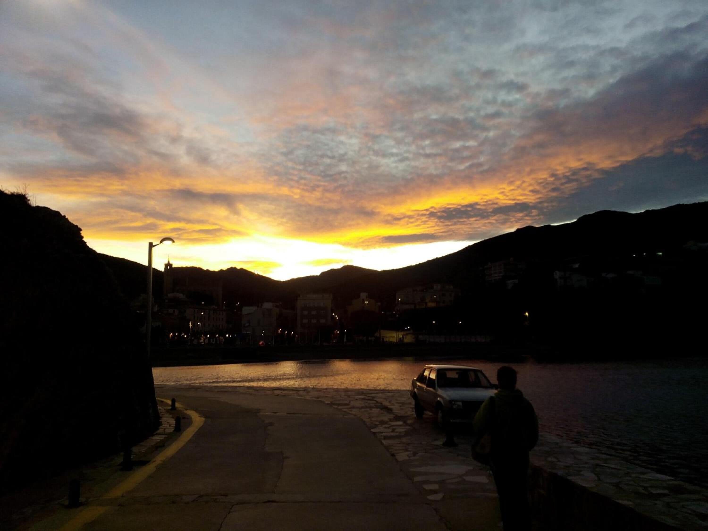 A sunset view with a body of water, a road, and buildings in the background.