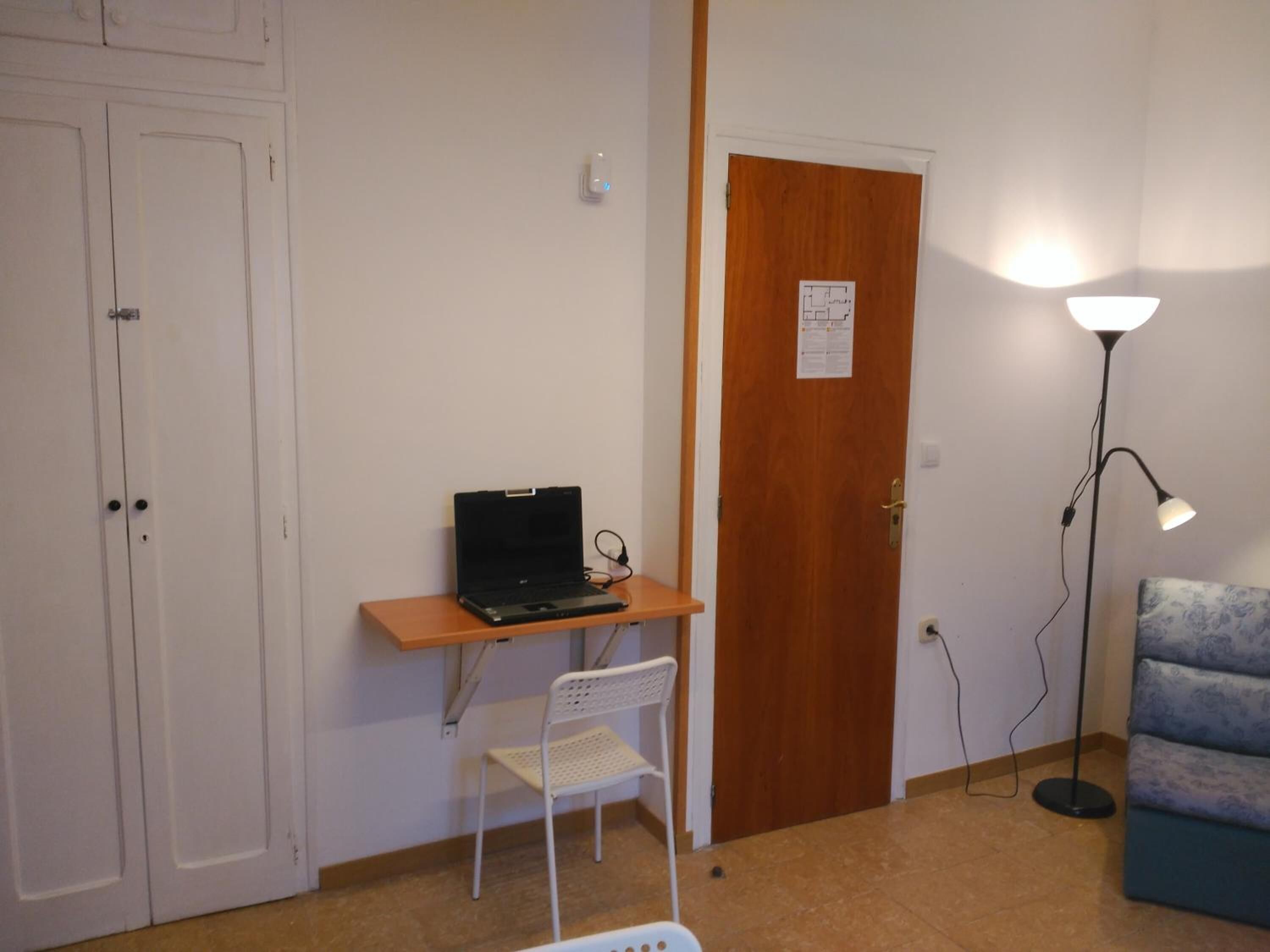 A room with a wooden desk, a laptop, a white chair, a blue patterned sofa, and a floor lamp.