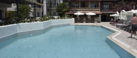 Una piscina con borde de azulejos, rodeada de sillones de descanso y sombrillas, junto a un edificio con balcones y paneles solares.