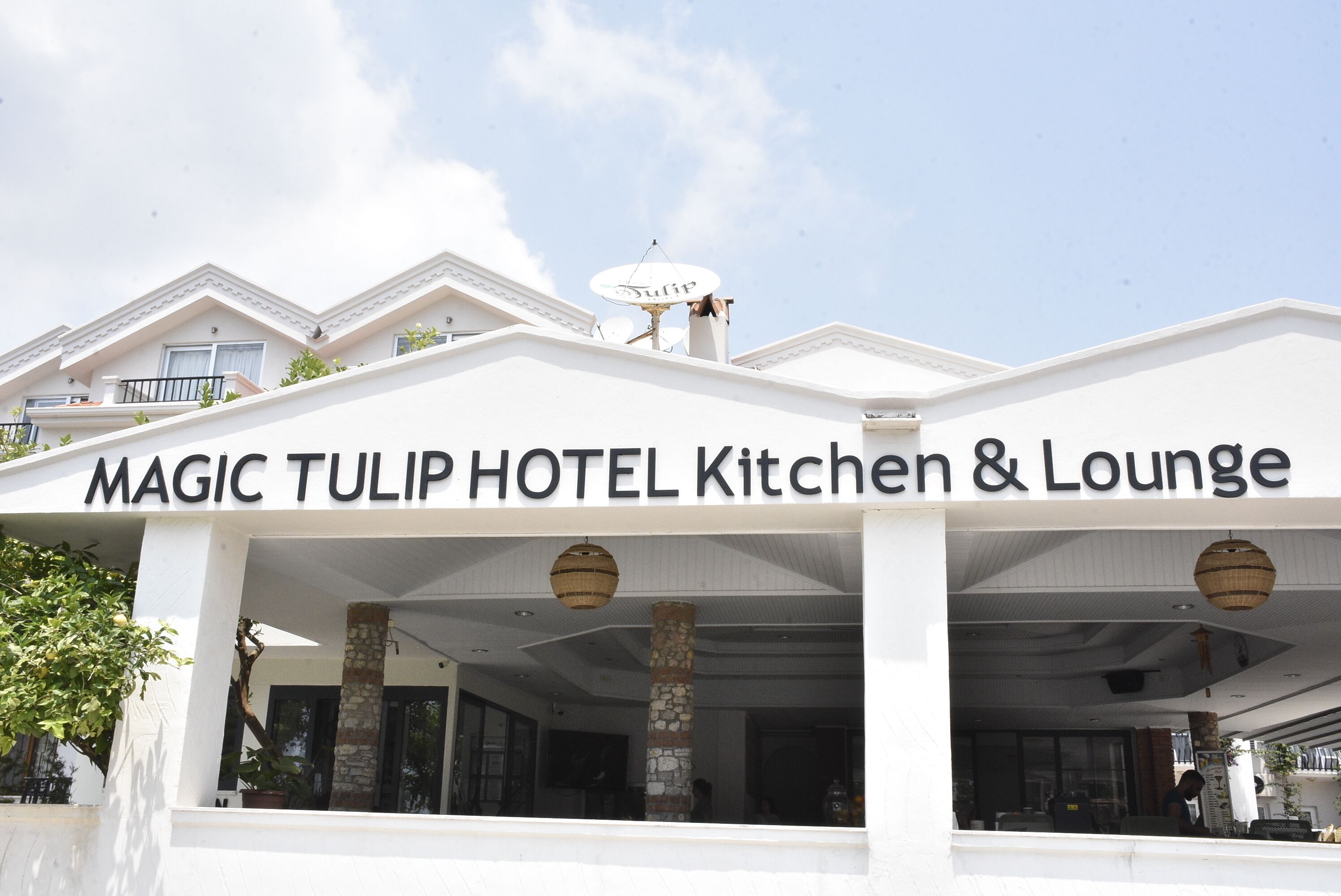 The exterior of the MAGIC TULIP HOTEL Kitchen & Lounge with a clear sign, stone pillars, and a satellite dish.