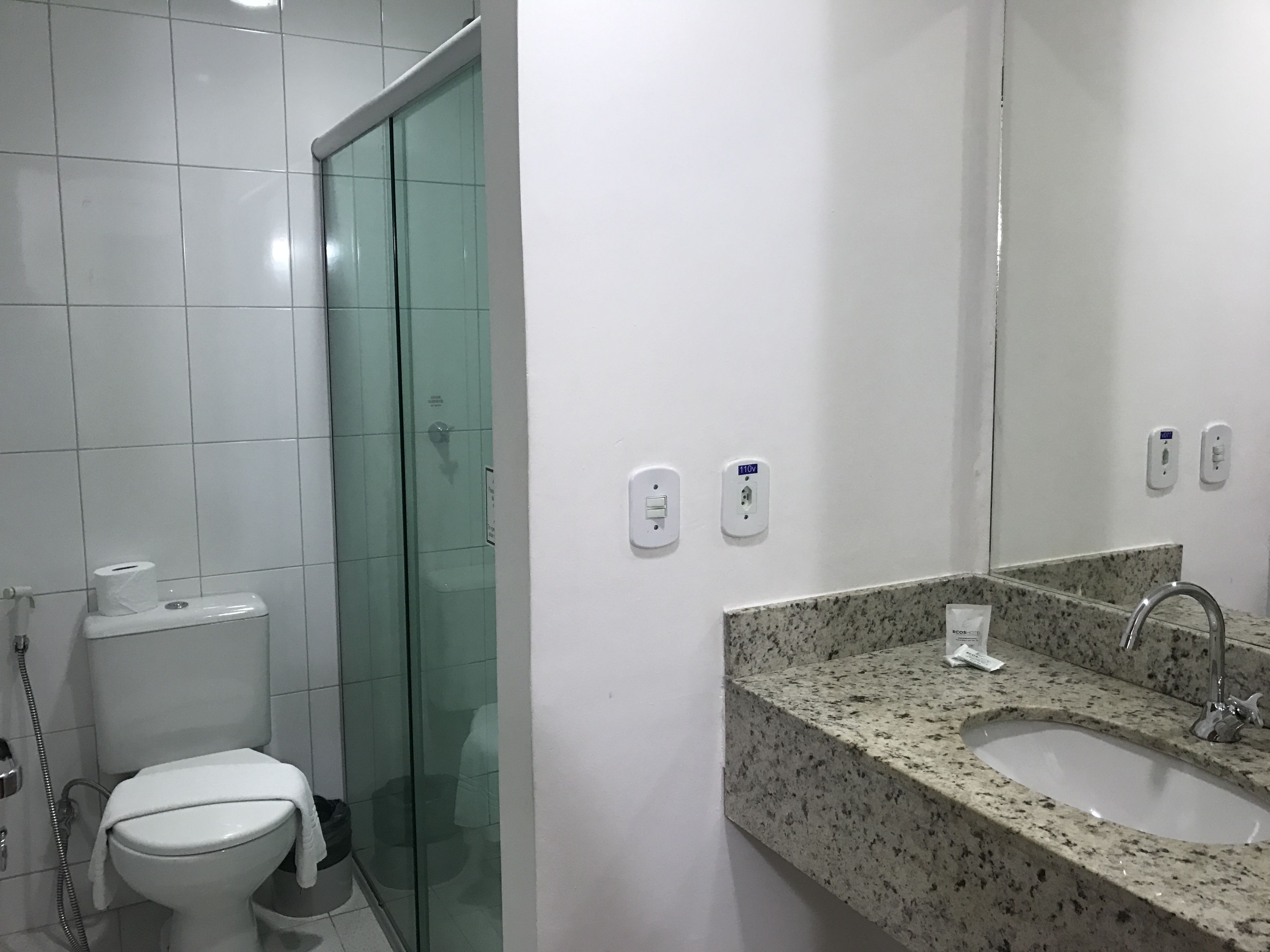 Un baño con inodoro, cabina de ducha de vidrio, lavamanos con encimera de granito y espejo.