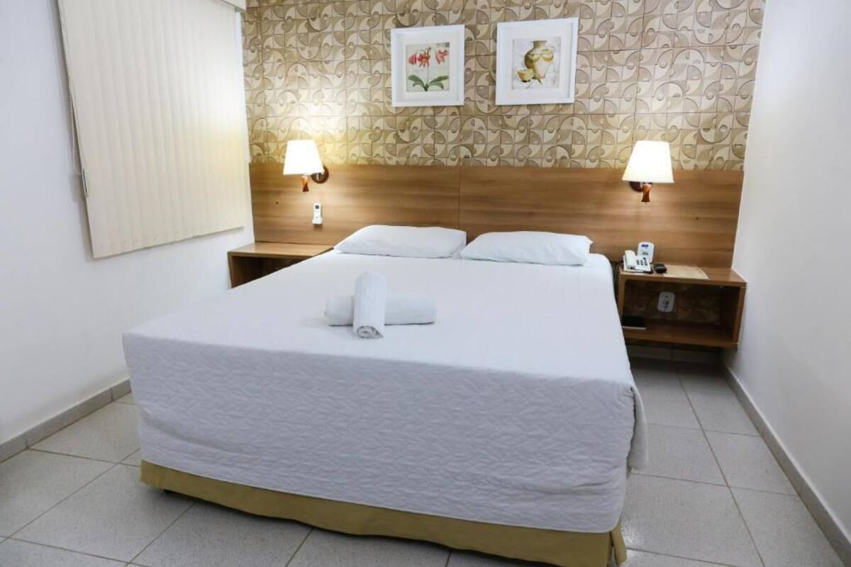 Ein ordentlich gemachtes Bett mit weißen Leinen und einem aufgerollten Handtuch, zwei Nachttischlampen und einem Holz-Kopfstück.