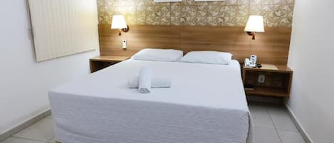 Een net opgemaakt bed met witte lakens en een opgerolde handdoek, twee bedlampjes en een houten hoofdbord.