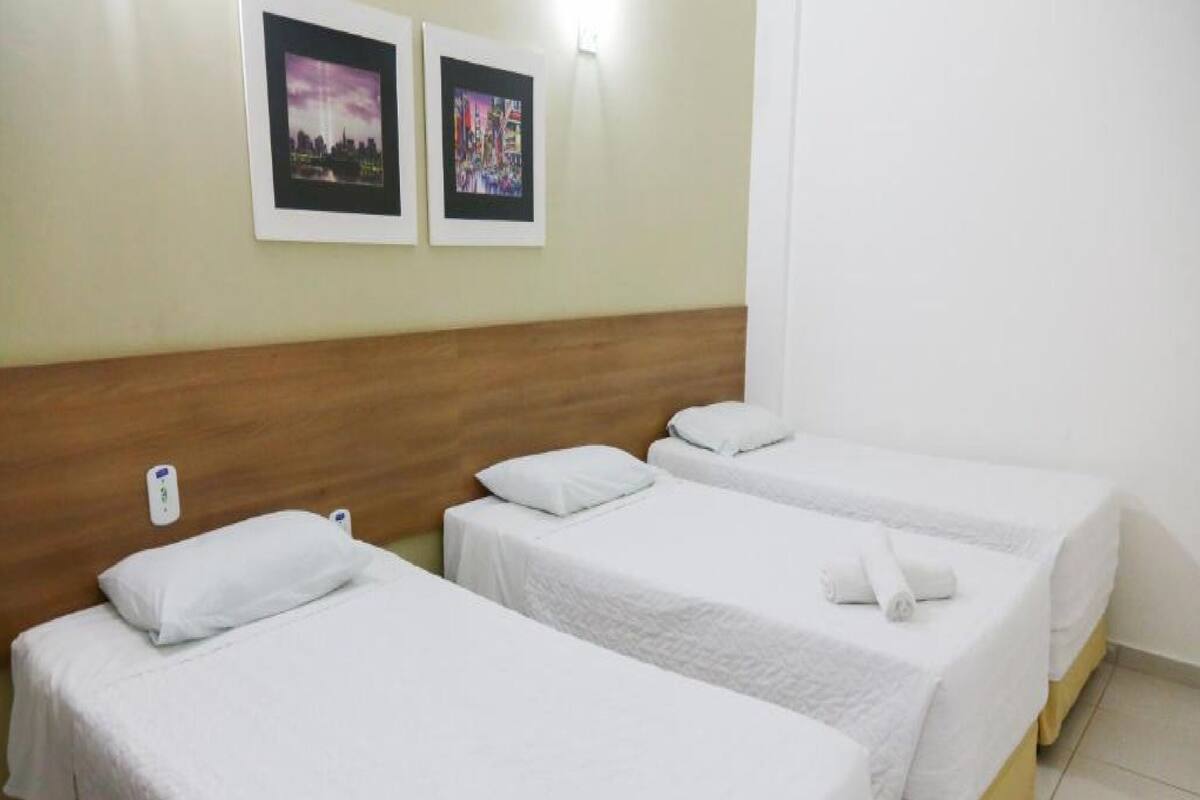 Ein Hotelzimmer mit drei Einzelbetten, jeweils mit weißer Bettwäsche und Kissen. An der Wand über den Betten hängen zwei gerahmte Bilder.