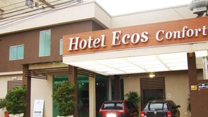 A hotel entrance with a sign reading "Hotel Ecos Comfort". There are two cars parked in front, and the building has a modern design with large windows and a covered entrance. - Ecos Conforto (Porto Velho)
