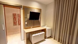 A modern bedroom with a flat-screen TV mounted above a wooden desk, a small bedside table, and a storage bench. - Ecos Conforto (Porto Velho)