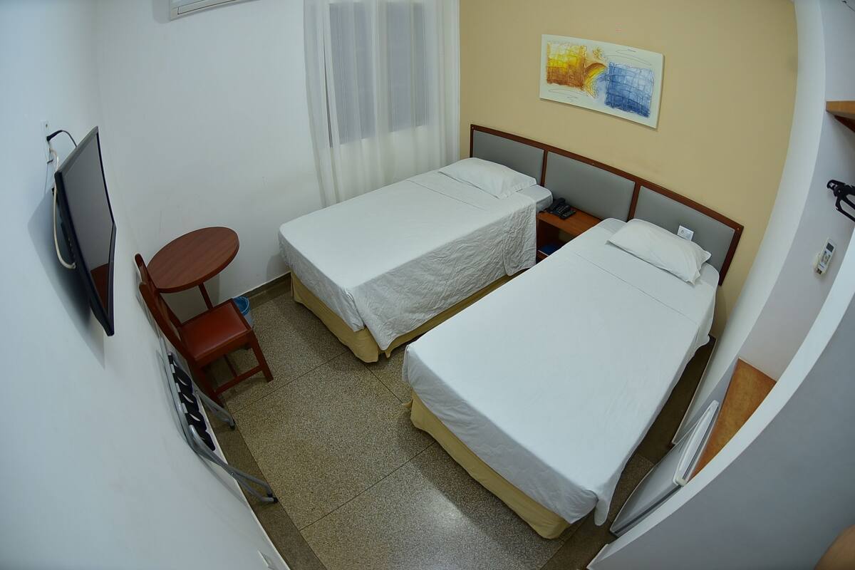 Habitació d'hotel petita amb dos llits individuals, una taula de treball, una cadira, un televisor i una finestra amb cortines.