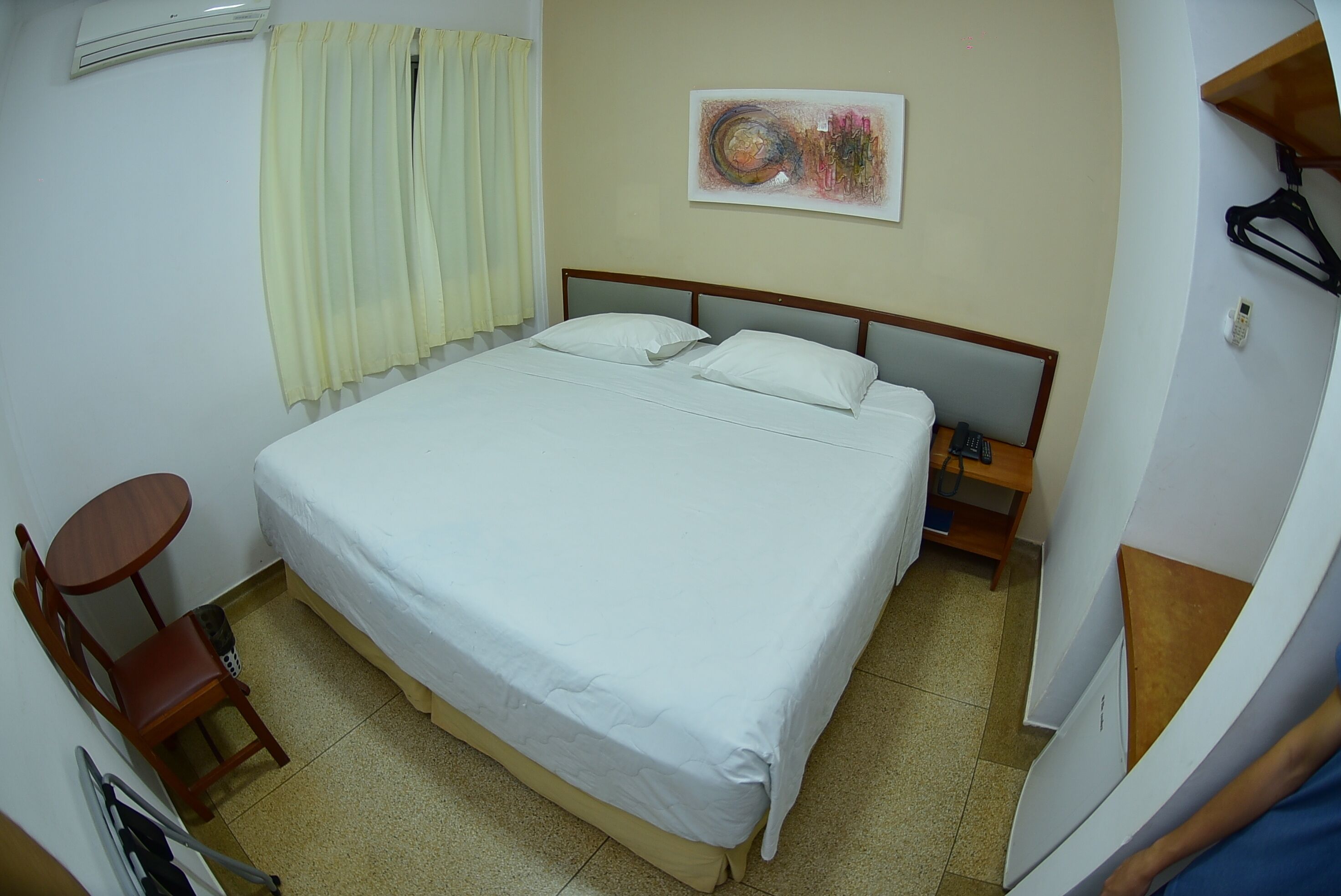 A hotel room with a large bed, a small round table, a chair, a wall-mounted air conditioner, a painting, and a telephone.