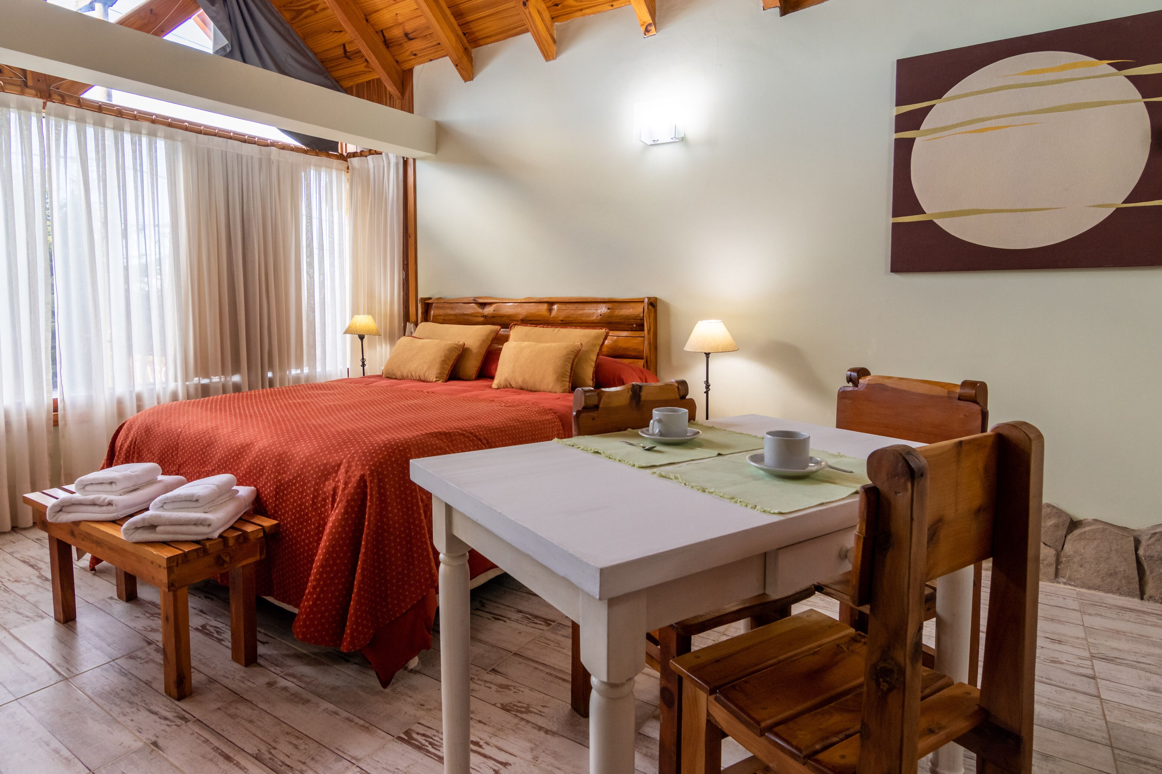 A hotel room with a wooden bed, a table with a cup, a chair, a bench with towels, and a large window with curtains.