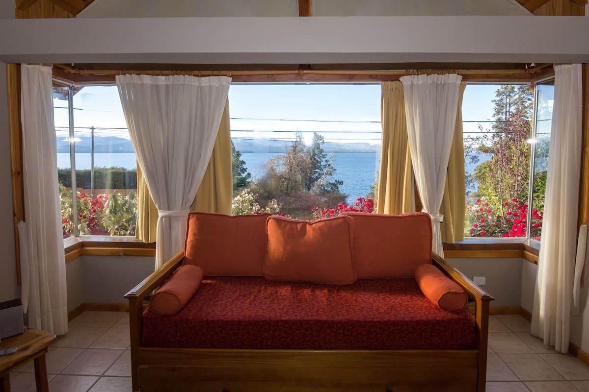 Quarto com cama de madeira, janelas amplas com cortinas e vista para um lago e árvores.