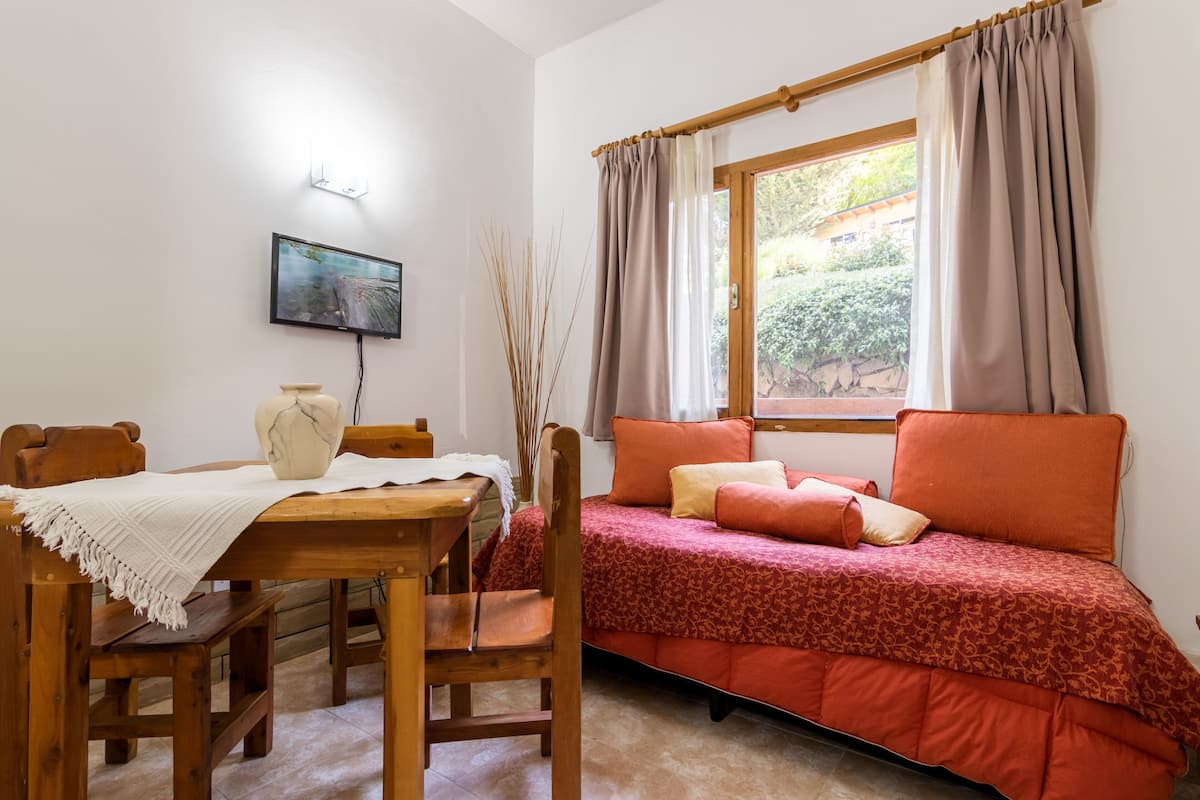 Um quarto aconchegante com uma mesa de madeira, cadeiras, uma cama com roupa de cama vermelha, uma janela com cortinas e uma televisão fixada na parede.