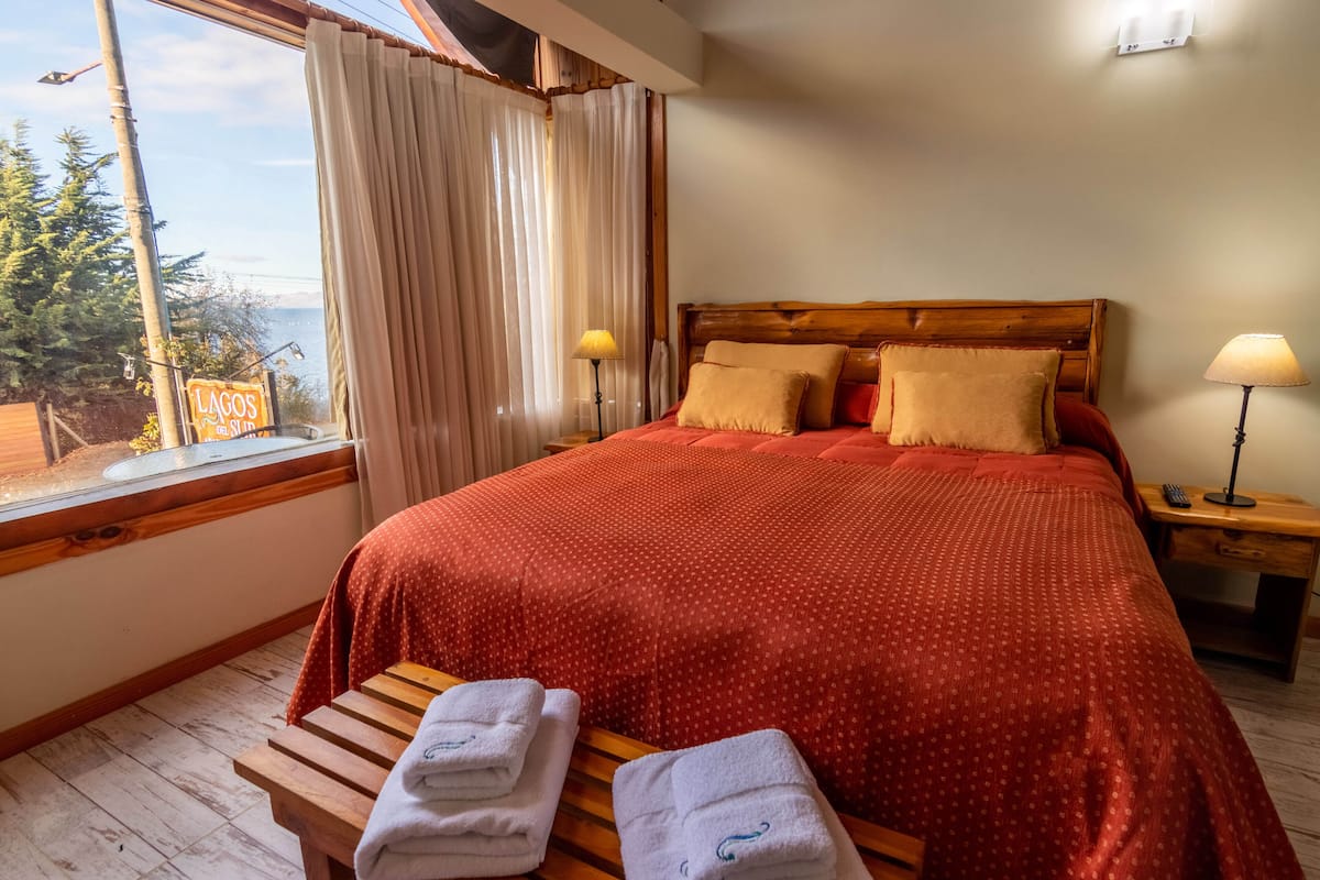 Quarto com cama grande, cabeceira de madeira e vista para o exterior pela janela.
