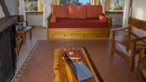 Un salon chaleureux avec un plafond en bois, un canapé rouge, une table en bois sur laquelle se trouve un ordinateur portable, et de grandes fenêtres offrant une vue sur la mer.