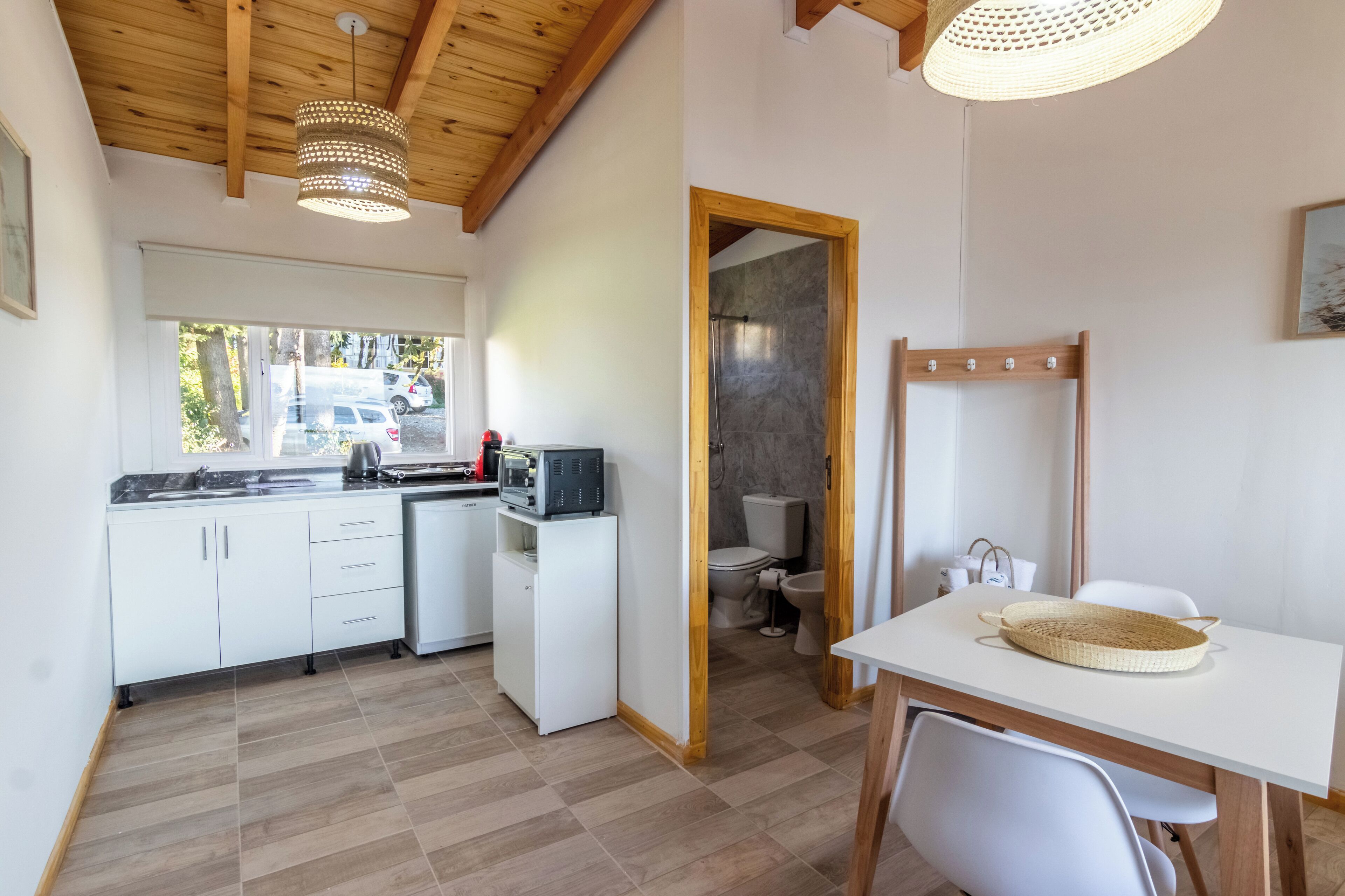 A compact kitchen with white cabinets, a microwave, and a sink. There's a dining table with chairs and a woven basket. A shower area is visible through an open door.
