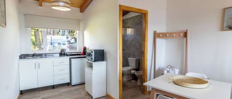 A compact kitchen with white cabinets, a microwave, and a sink. There's a dining table with chairs and a woven basket. A shower area is visible through an open door.