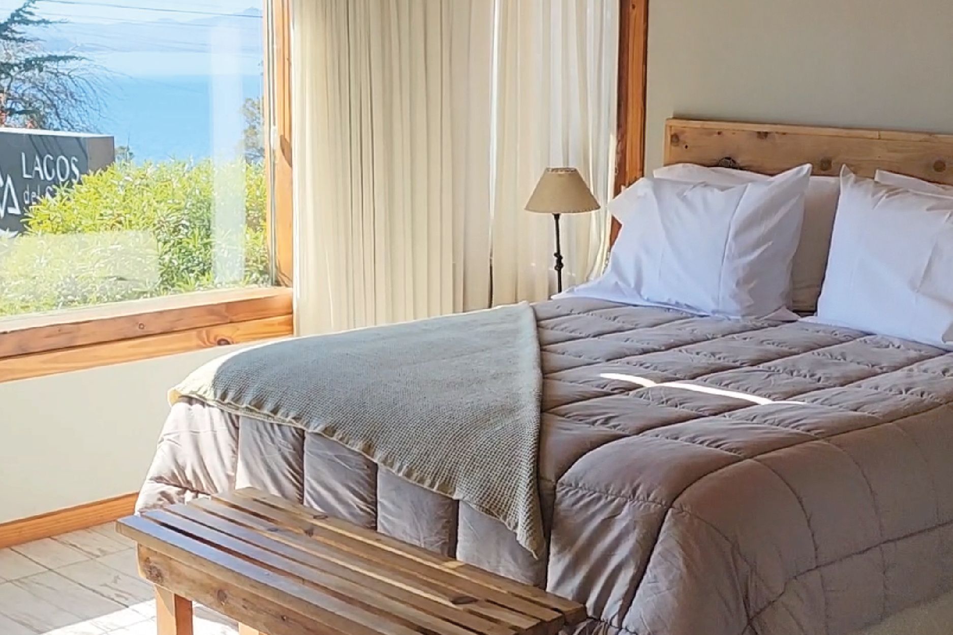 A bedroom with a bed, a bench, a lamp, and a view of the outdoors.