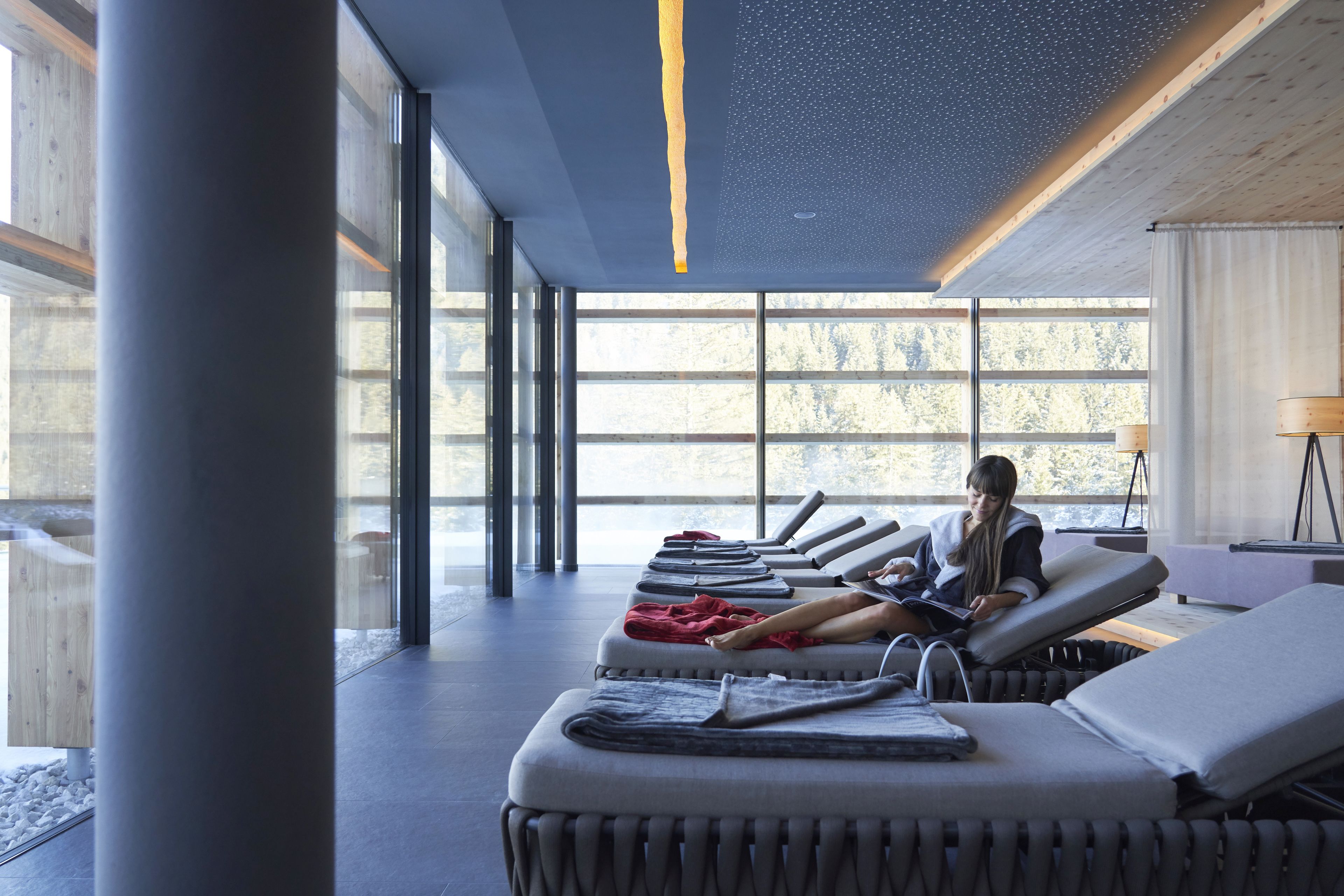 A modern spa room with reclining chairs, a large window with a view of trees, and a ceiling with a unique light fixture.