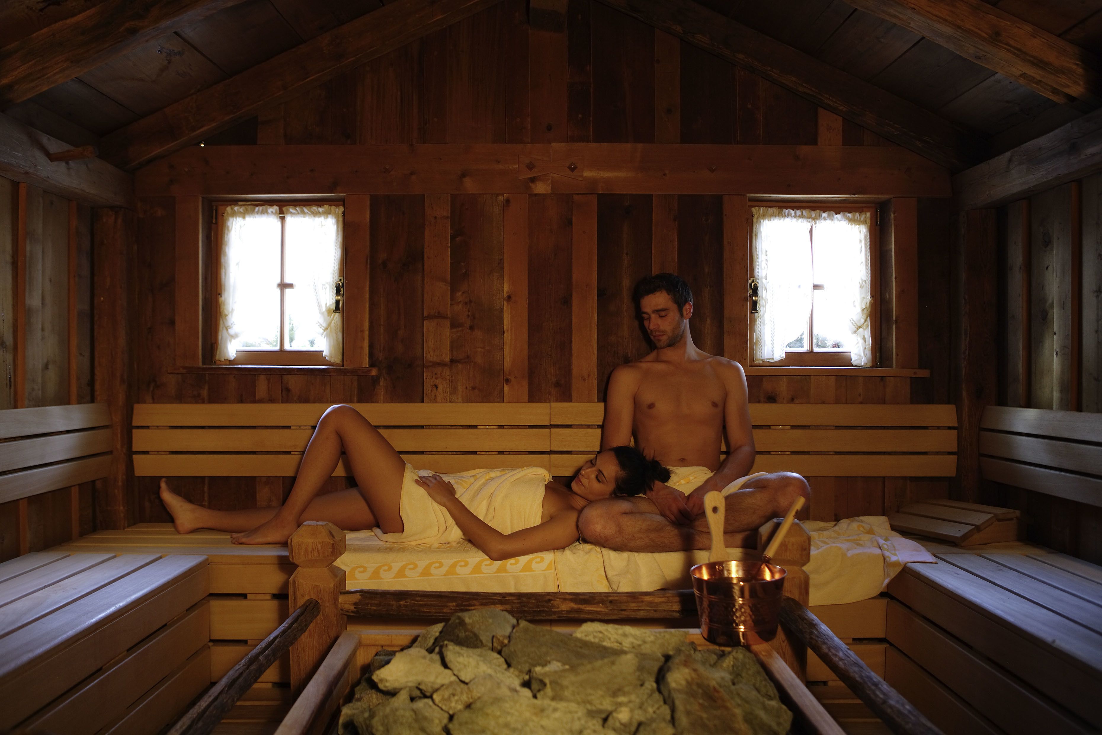 A wooden cabin with a wooden bed, two windows, and a stone fireplace.