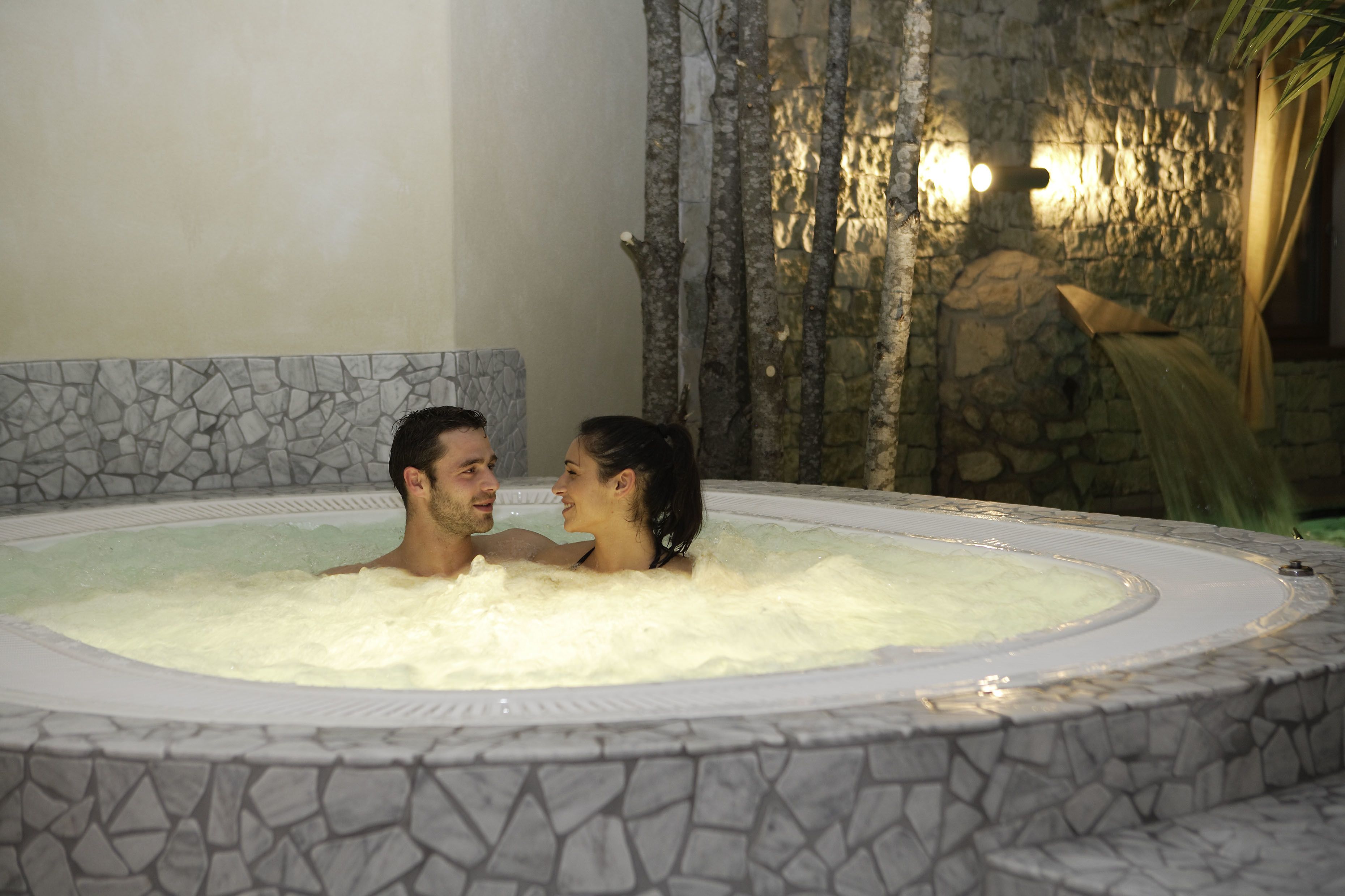 A couple in a stone-tubbed hot tub with a stone wall and a waterfall feature in the background.