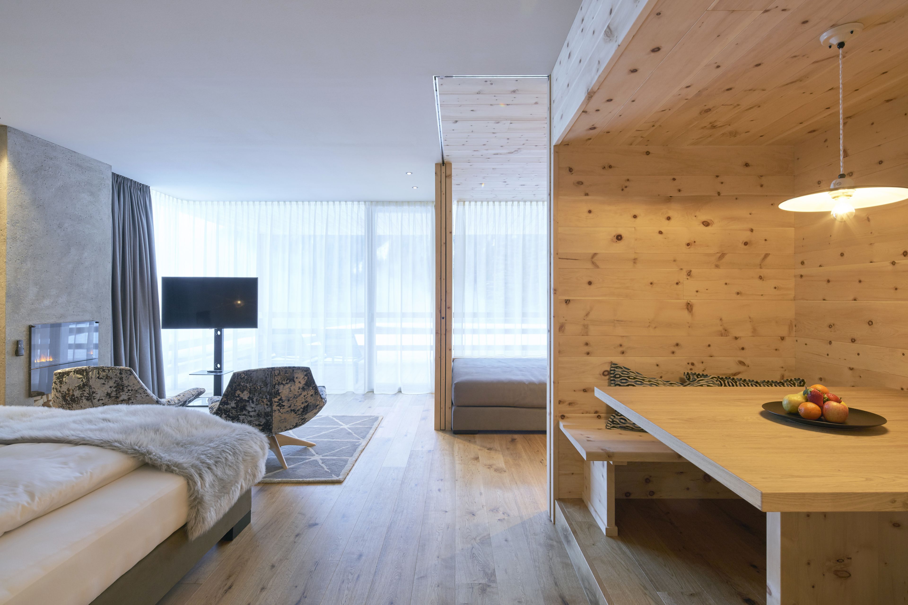 Una habitación de hotel moderna con una cama, un televisor, una mesa de comedor con fruta y vistas a la ciudad a través de grandes ventanales.