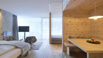 Una habitación de hotel moderna con una cama, un televisor, una mesa de comedor con fruta y vistas a la ciudad a través de grandes ventanales.