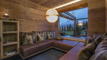 Interior acogedor de una cabaña con detalles de madera, un ventanal con vistas a árboles, una zona de estar con cojines y una estantería.