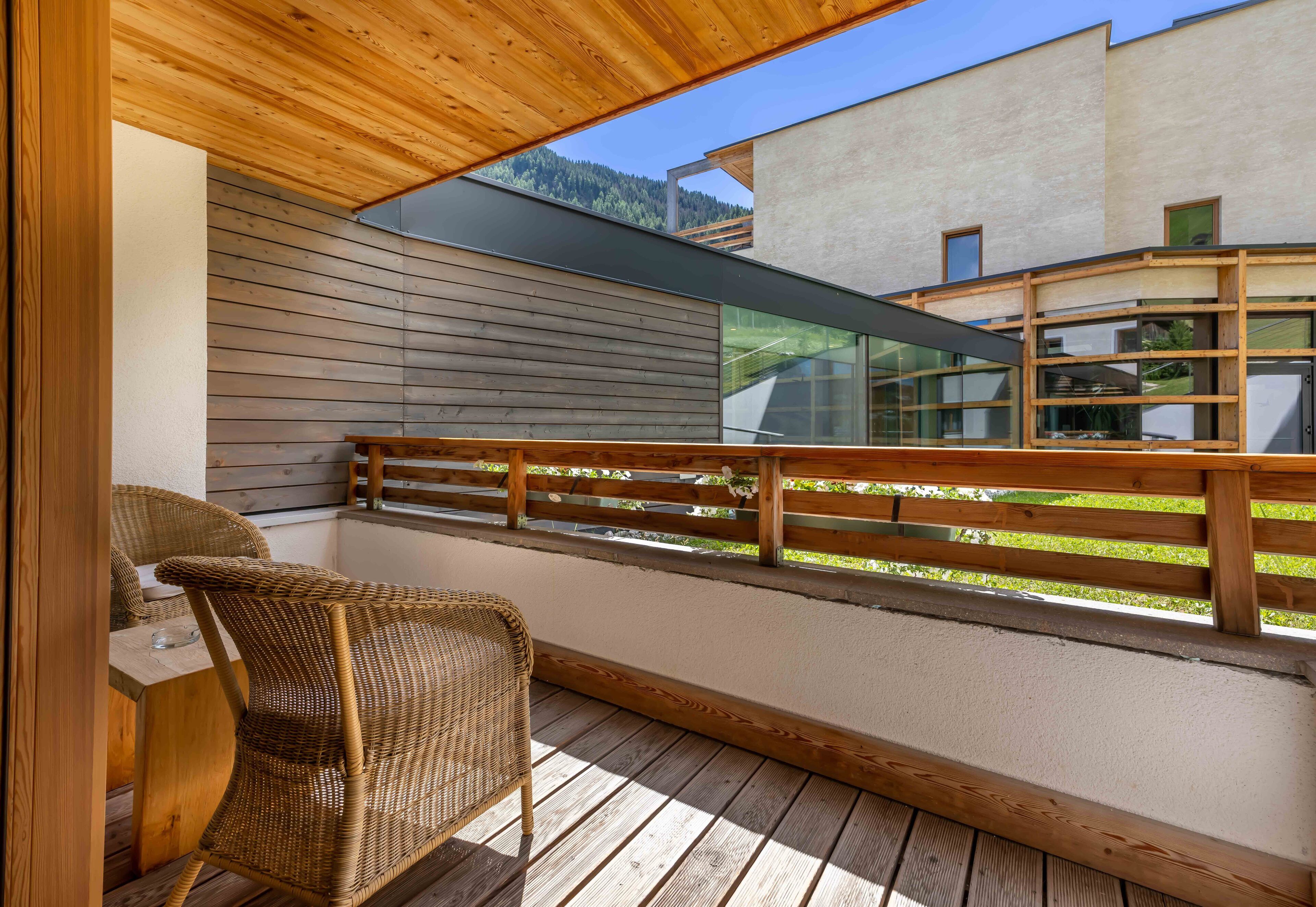 A wooden deck with a wicker chair and a table, overlooking a modern building and a forested hillside.