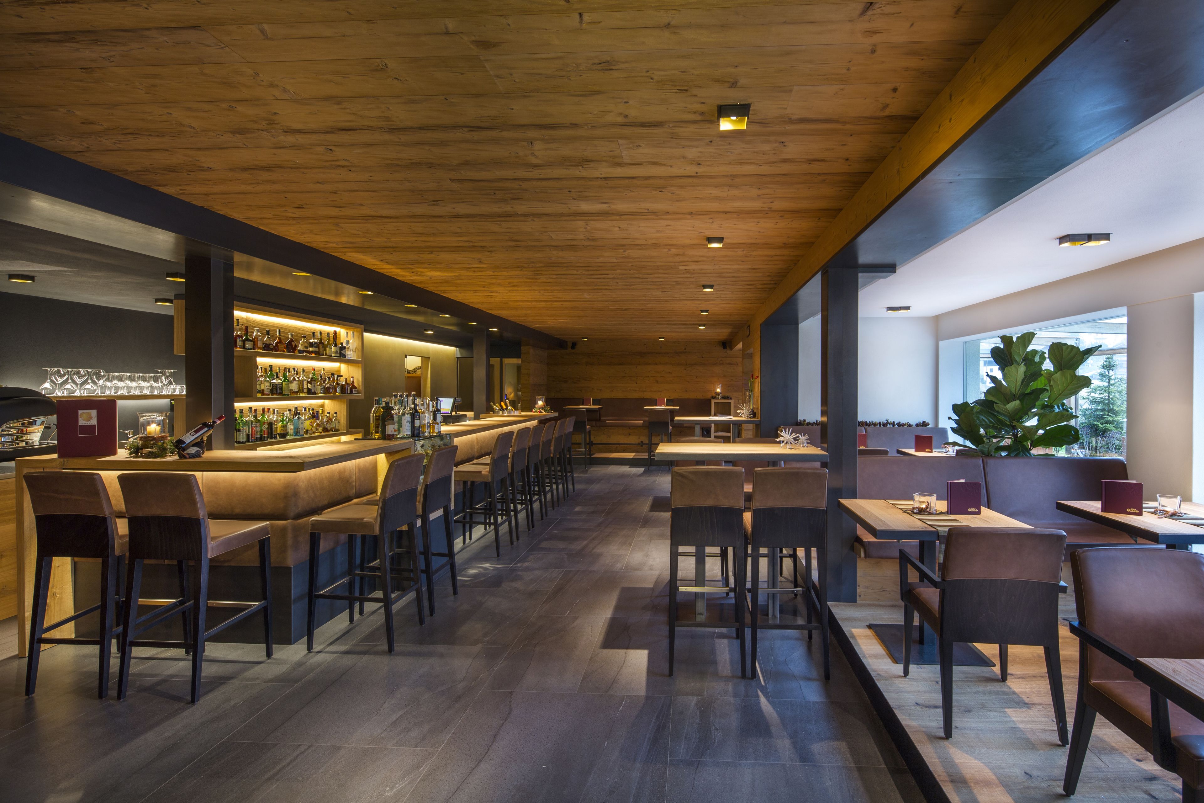 A modern bar area with a long counter, multiple seating options, and a large window providing natural light.