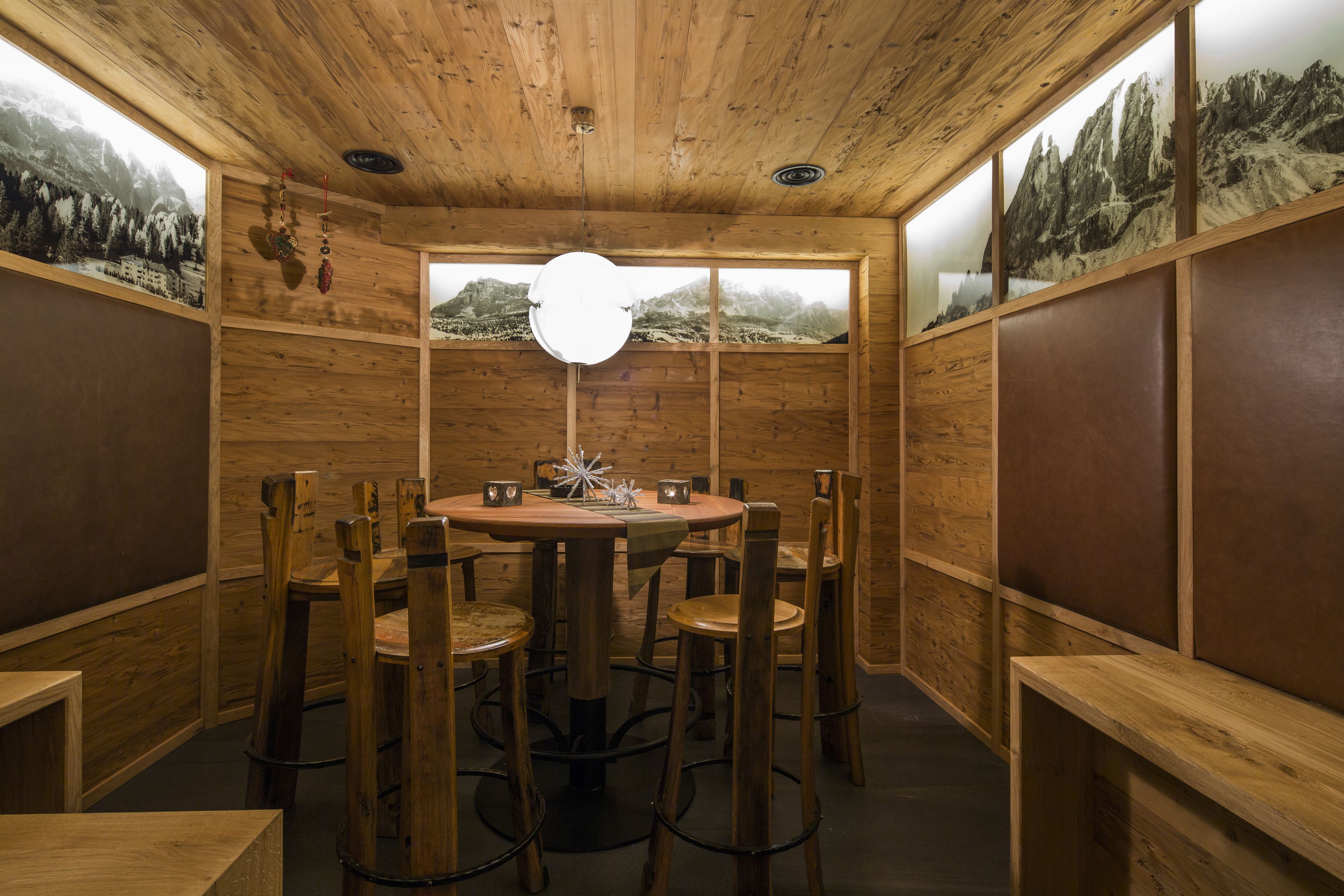 A wooden interior with a round table and chairs, wooden walls, and a large window with a mountain view.