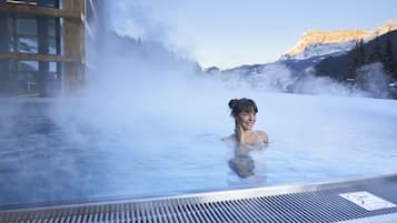 Uma pessoa em uma nascente termal com vapor subindo, rodeada por um edifício de madeira e montanhas cobertas de neve ao fundo.