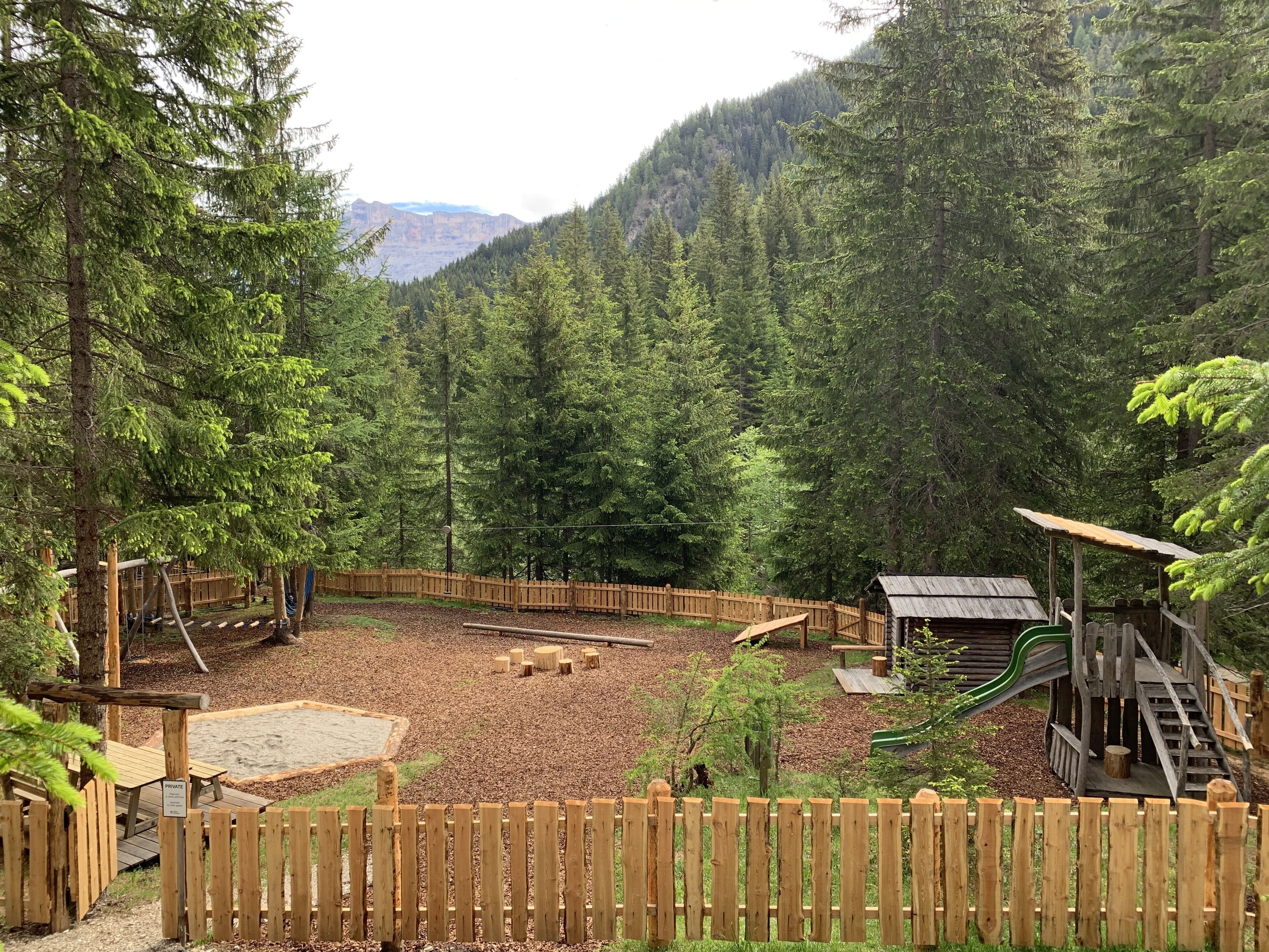 Un patio de juegos con estructuras de madera y un área cercada, rodeado de un bosque denso y montañas al fondo.
