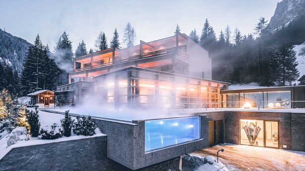 A modern multi-story building with large glass windows, surrounded by snow-covered trees and mountains.