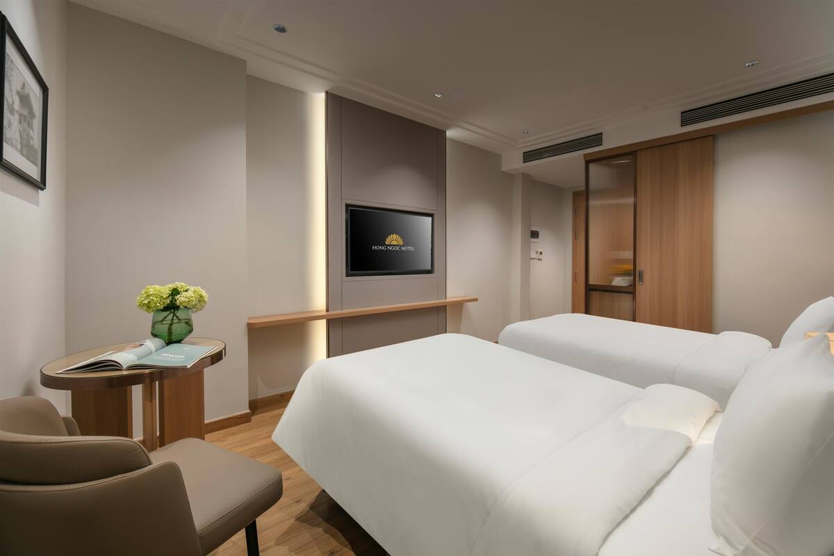 A hotel room with two beds, a TV, a desk with a vase of flowers, and a framed picture on the wall.