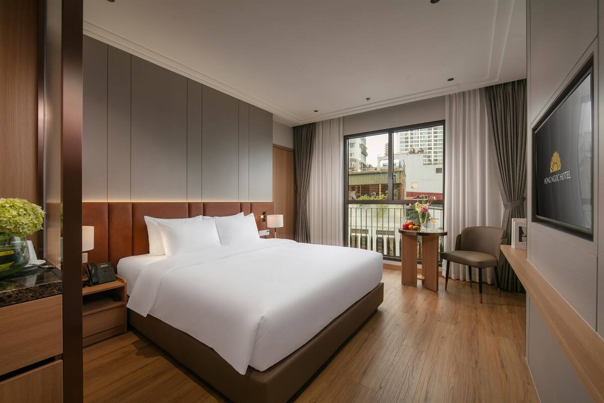A hotel room with a large bed, a desk with a chair, a small table with flowers, and a TV on the wall.