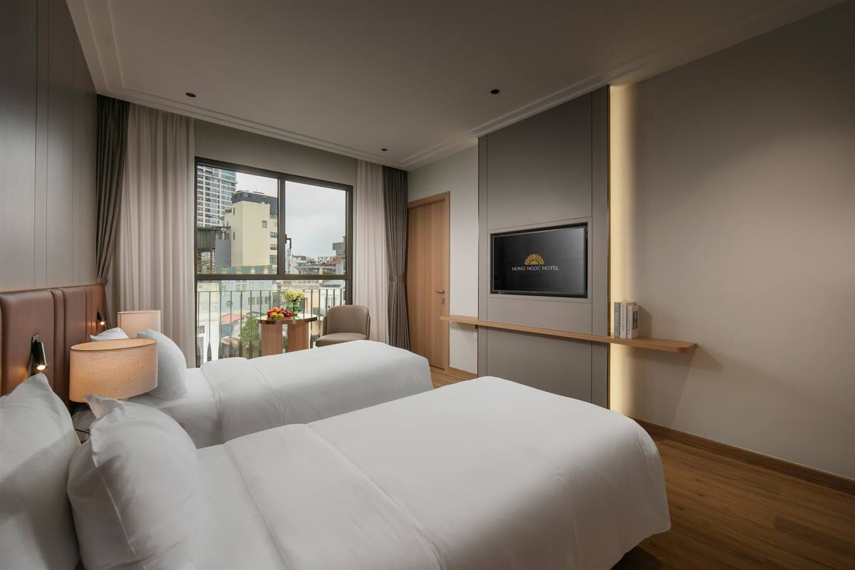 A hotel room with two beds, a TV, a window with curtains, a small table with a lamp, and a view of buildings outside.