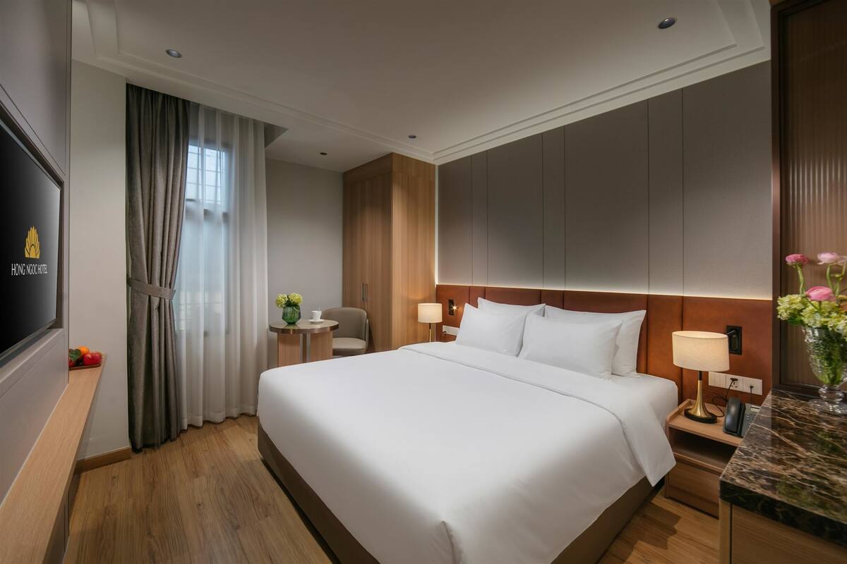 A hotel room with a large bed, two bedside tables with lamps, a TV, a small round table with a chair, and a window with curtains.