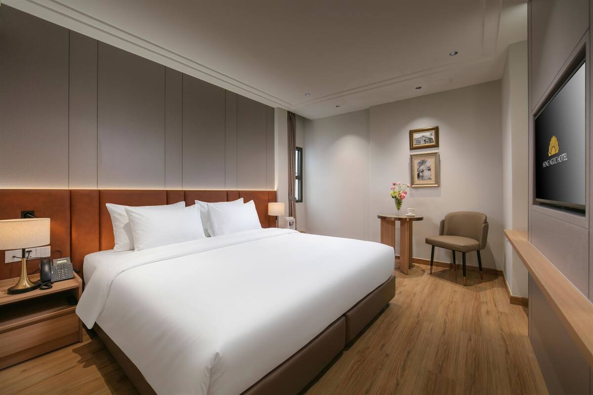 A hotel room with a large bed, bedside table with a lamp, a small table with a vase, a chair, and a TV mounted on the wall.
