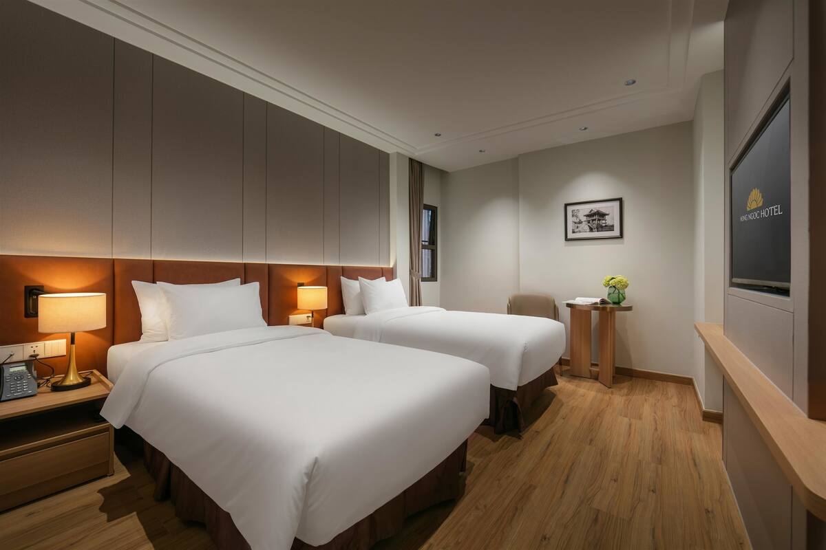 A hotel room with two beds, a bedside table with a lamp, a small table with a vase, and a framed picture on the wall.