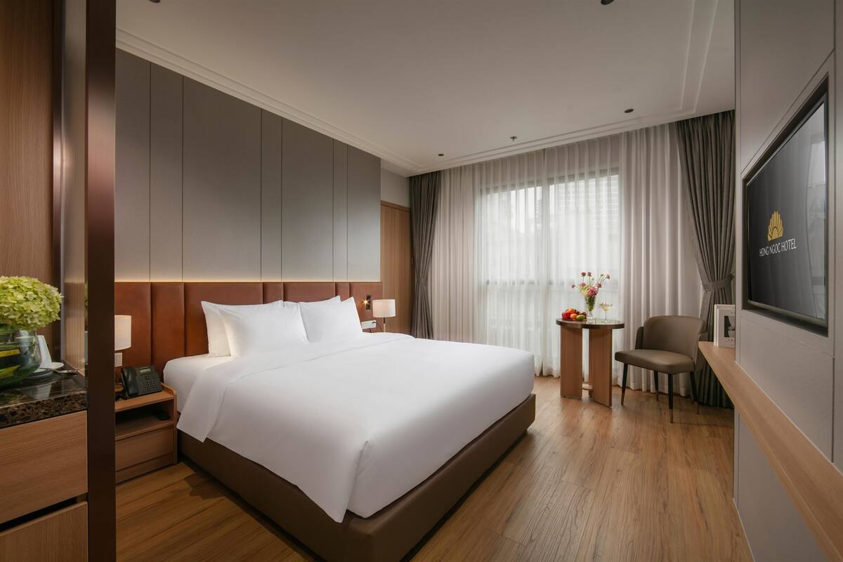 A hotel room with a large bed, a bedside table with a lamp, a small table with flowers, a chair, and a TV mounted on the wall.